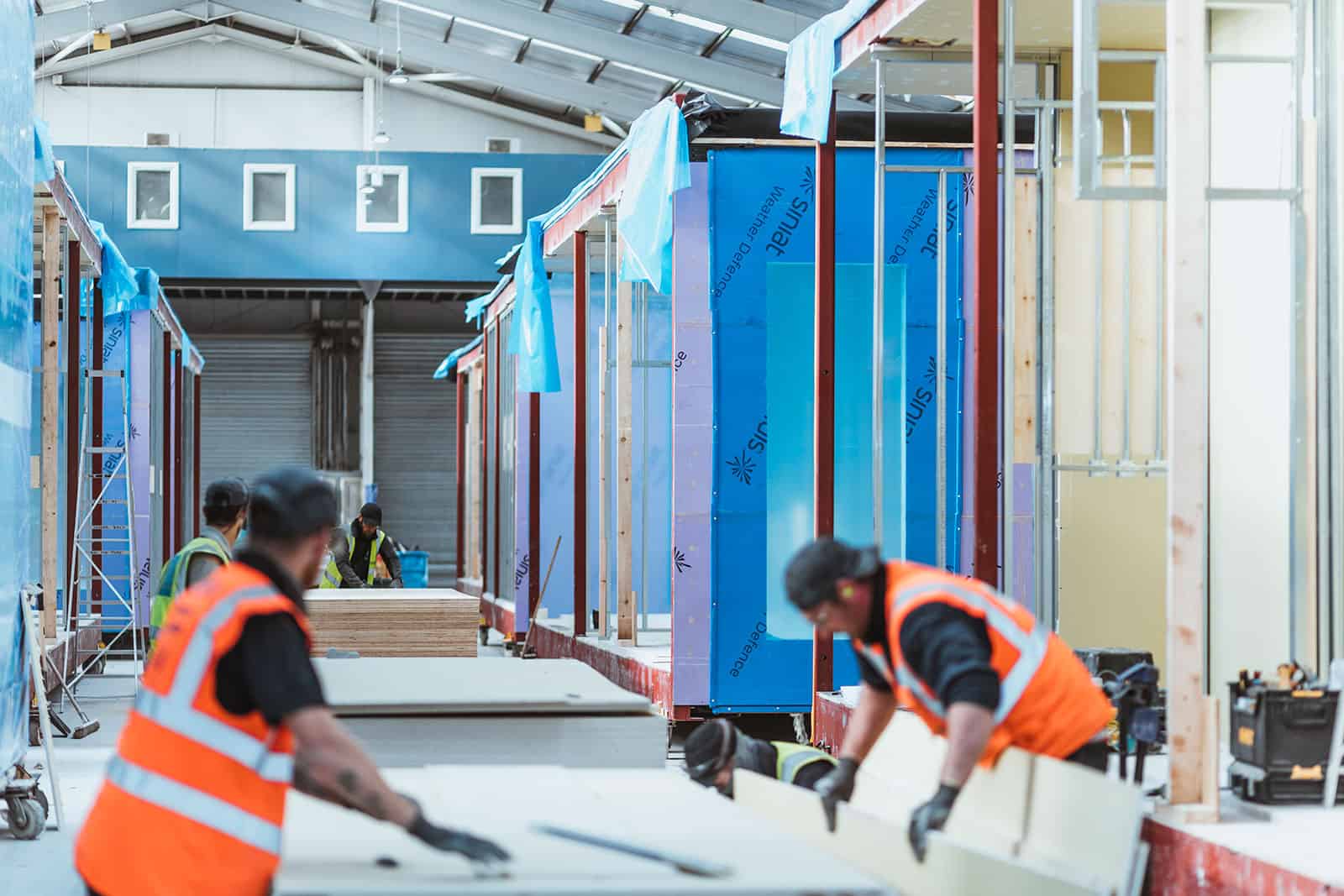 Construction workers building units of a modular building in a warehouse, off-site so that it can be taken to the actual site and constructed there. 