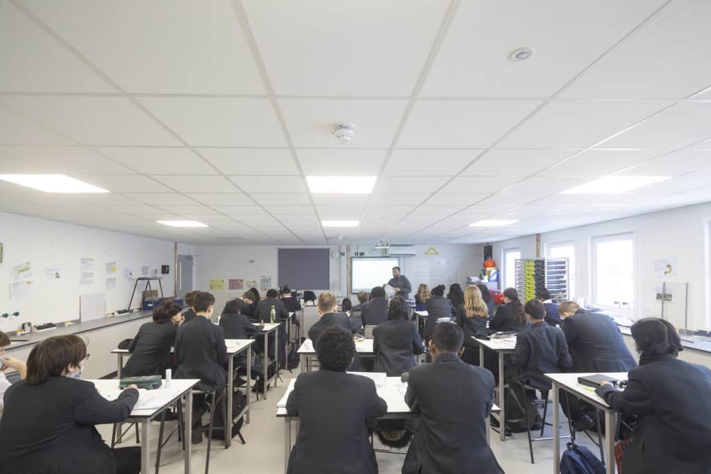 Inside Temporary classroom, filled with students wearing black blazers, sat at desks. Teacher at the front of the room standing next to the interactive whiteboard.