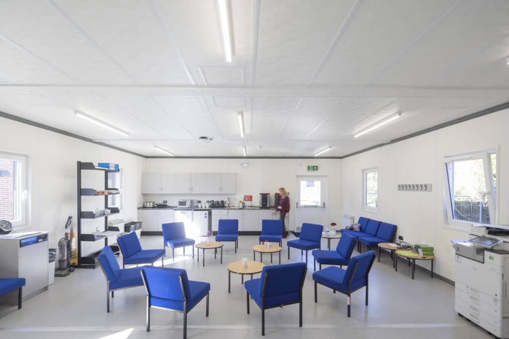 Inside a modular classroom, white walls and ceilings, 3 ables in the centre of the room, surrounded by blue chairs. 