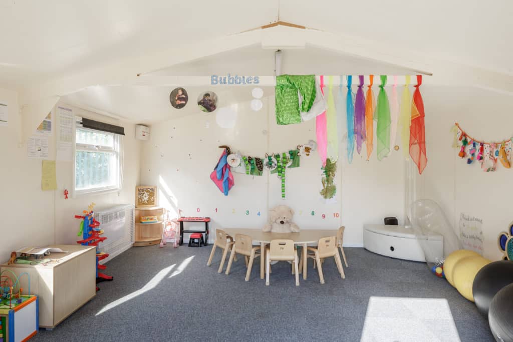 Inside temporary classroom for young children. Very small chairs and desks, with colourful resources on the walls and toys at the edges of the well-lit room. 