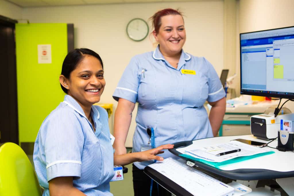 2 smiling nurses gathered around a desk with medical charts on and a computer, inside a modular healthcare unit. 