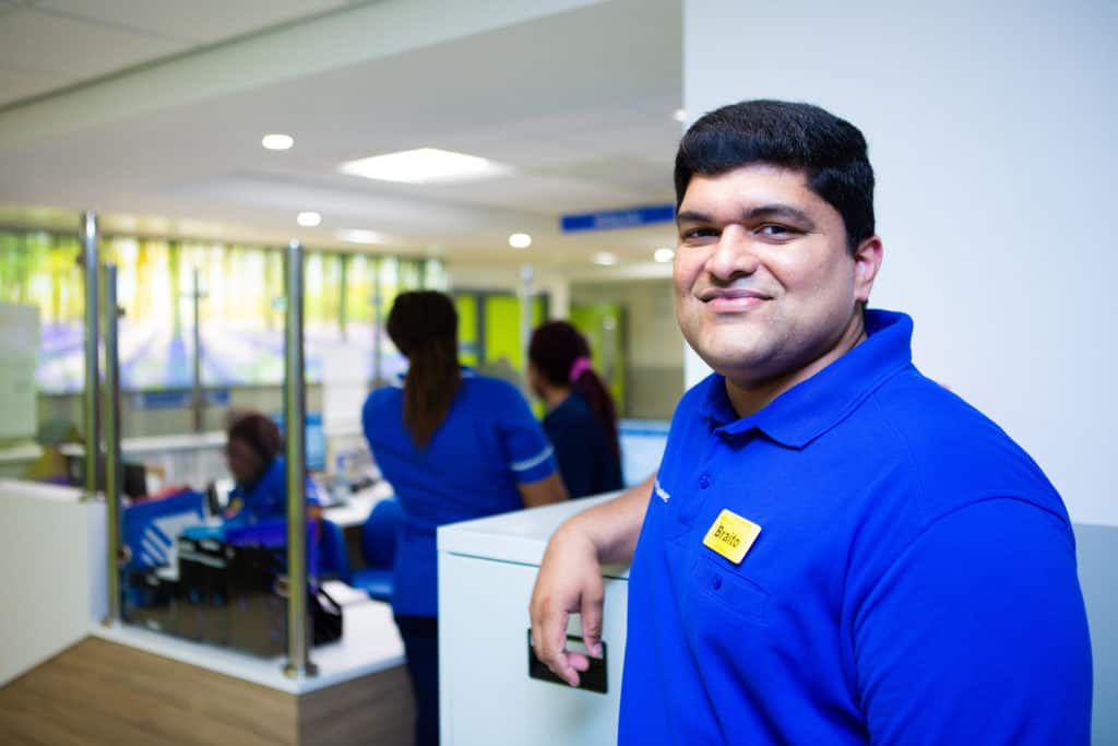 Male employee smiling, infront of female nurses gathered around a reception desk in modular hospital building.