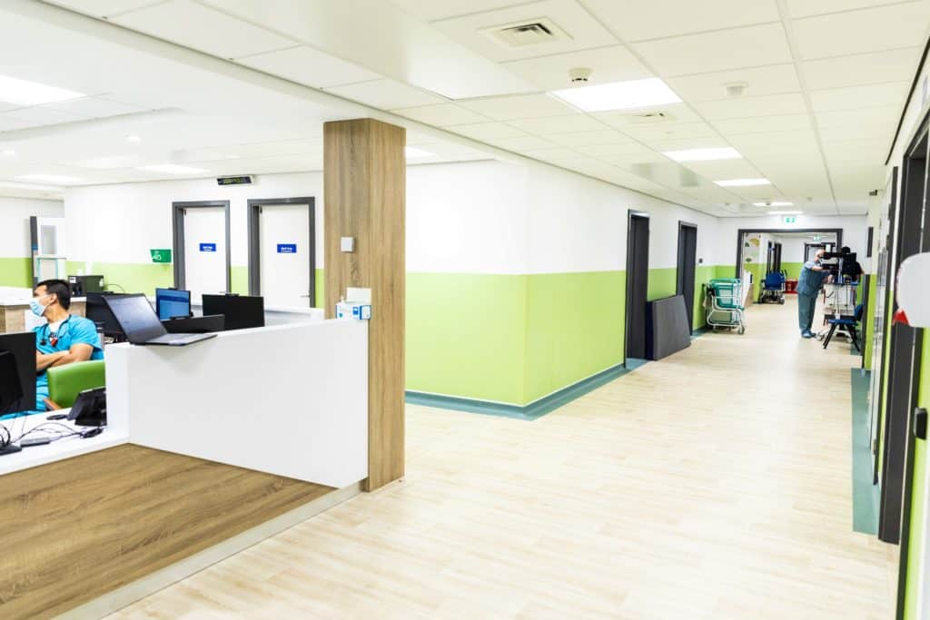 Inside view of a modular healthcare facility built for St Peter's Hospital. Long corridors with green and white walls and a male nurse sat at a reception desk wearing a mask. 