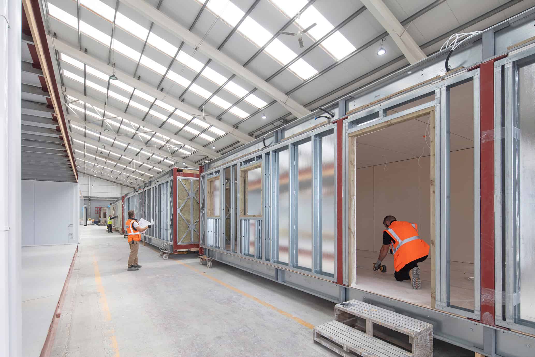 Construction workers working inside a nearly complete modular unit, in a warehouse. One man is reading the prefab designs outside of the units. 