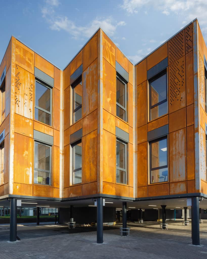 Outside view of Modular building coated in aluminium powder. Multiple modular classrooms, in a zigzag shape, onto podiums that leave room for car parking.