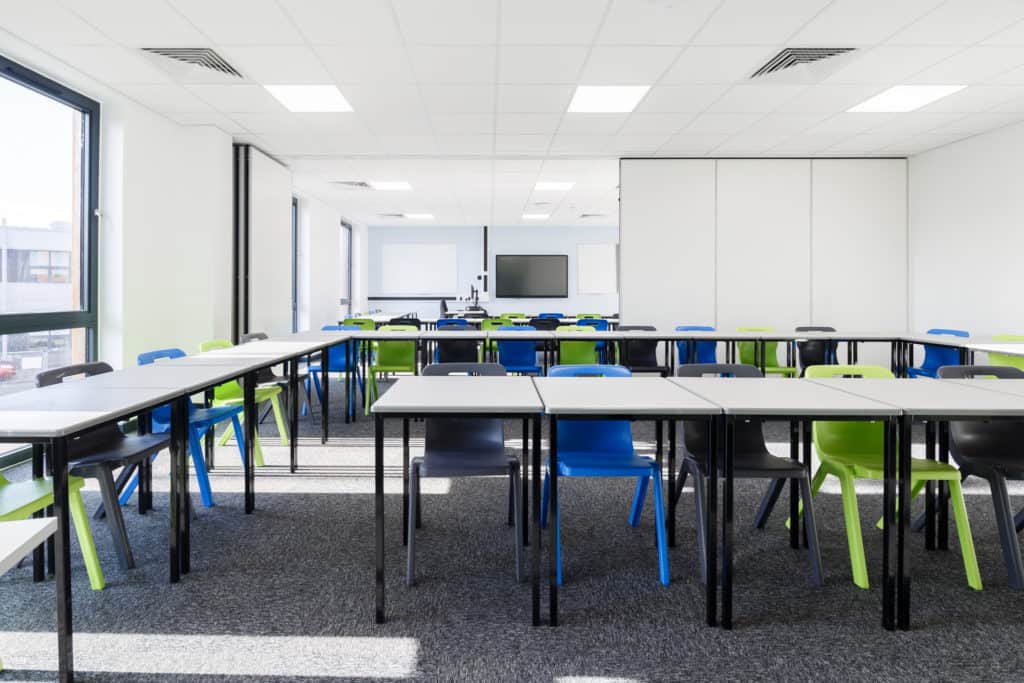 Modular classroom with white walls and several desks surrounded by black, green or blue chairs. Back of the classroom opens up, allowing to conjoin rooms.