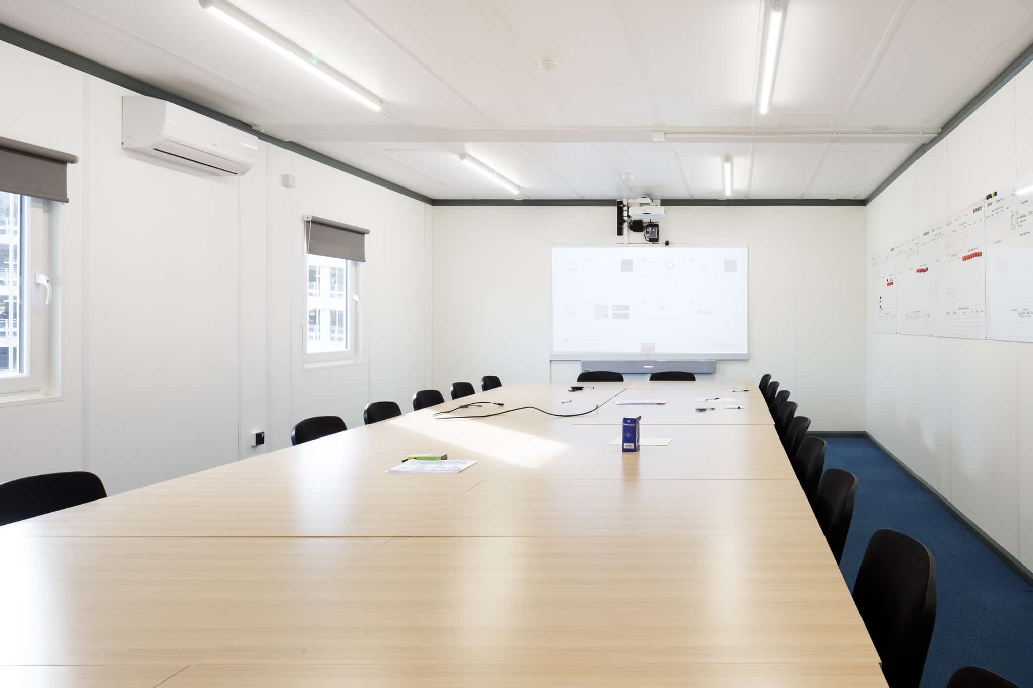 Inside temporary modular building. One room with a large table and several chairs surrounding. Along with a board and projector. 