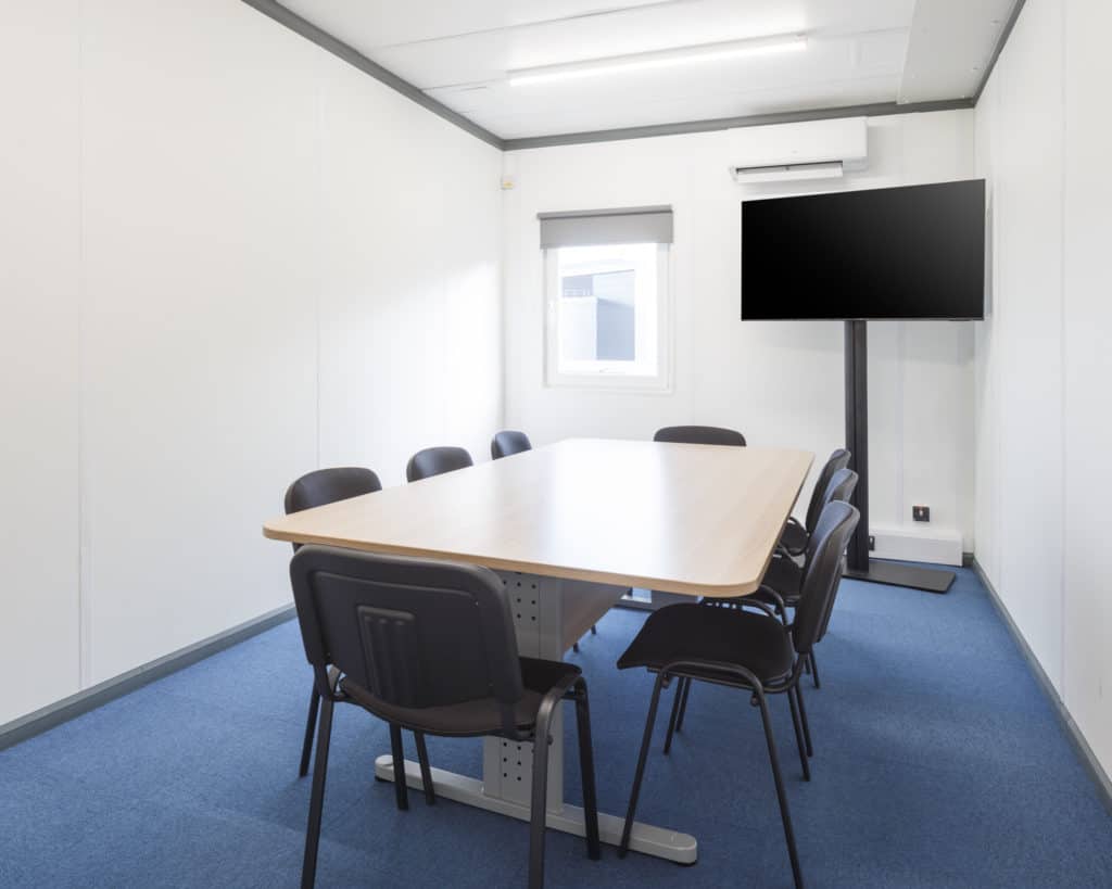 Inside temporary modular building. Table and chairs with a TV behind. White walls, blue carpet. 