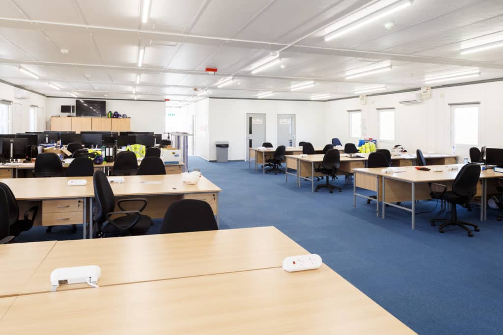 Inside view of modular building. Desks and chairs, windows, blue carpet, and white walls.