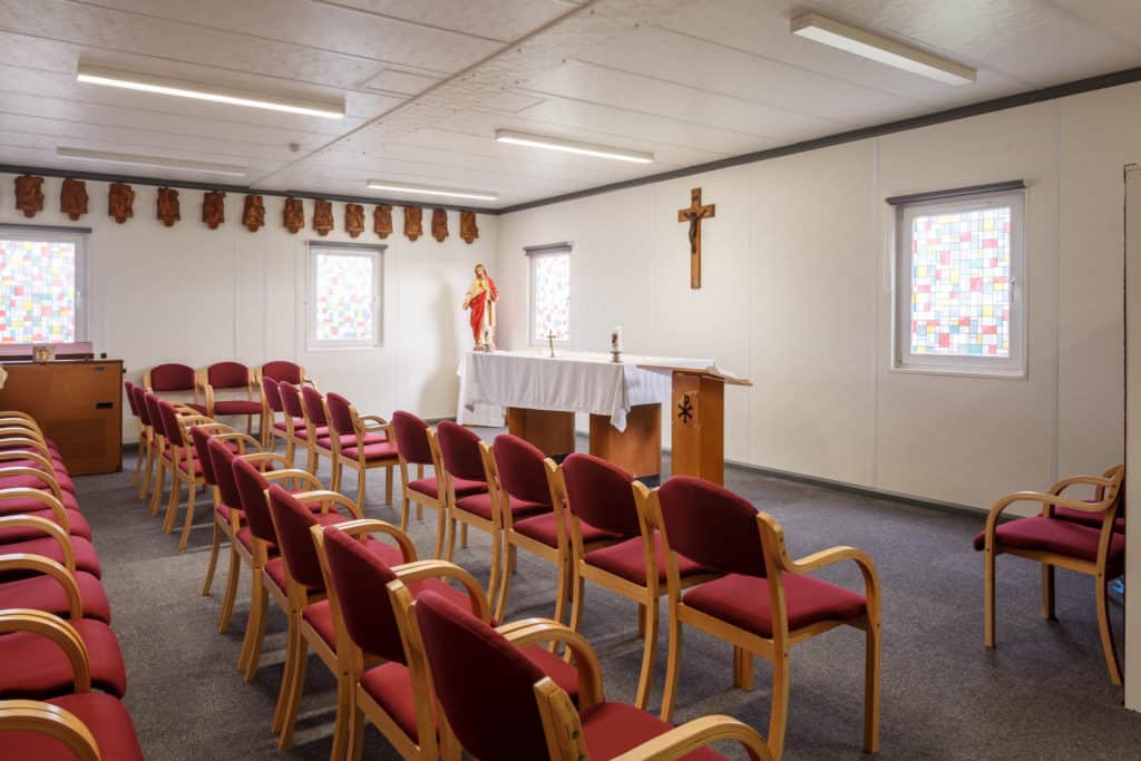 Church area inside the modular school. Cross on the wall. Chairs lined in front of a stand. Small stained glass windows.