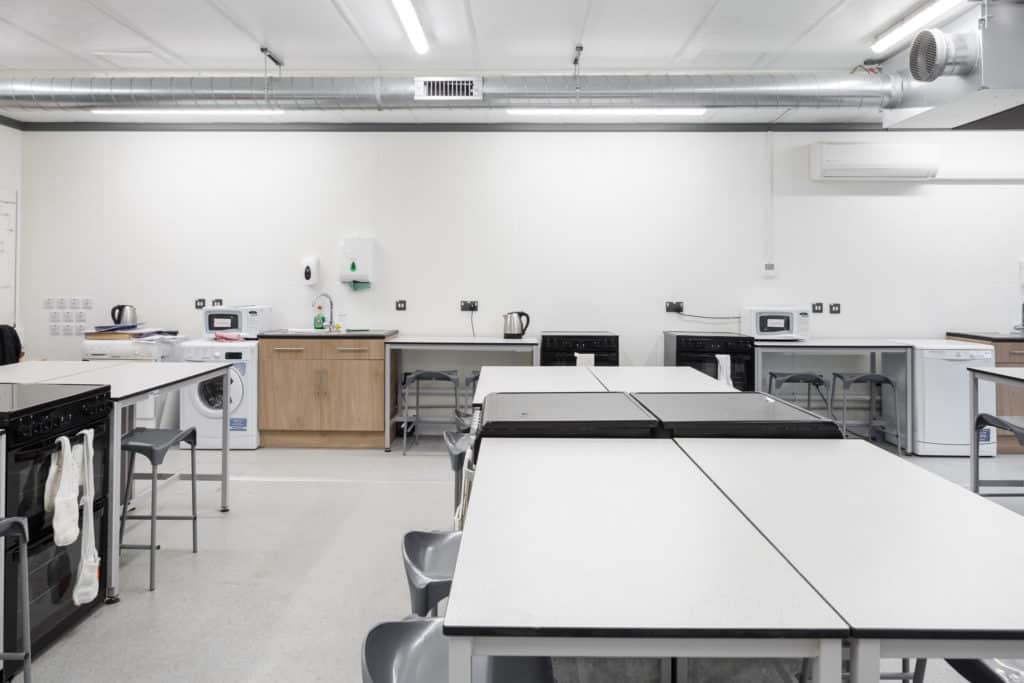 Inside the temporary classroom building. Long table with chairs, a washing machine, stools, sink and hand dryer. 