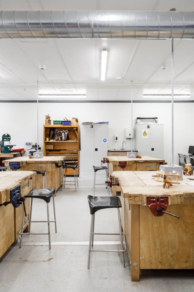 Temporary classroom interior, D&T area. Wooden work benches with clamps attached, and studen stools. Bright space, white ceilings and walls. 