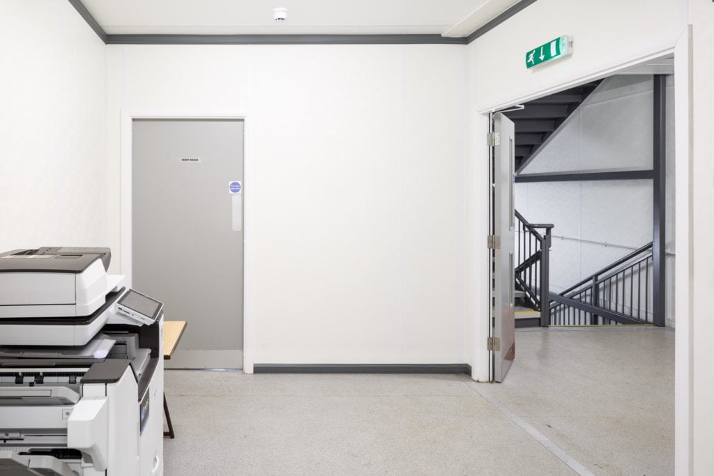 Large printer in a white room inside the modular school. Door and stair case in view.