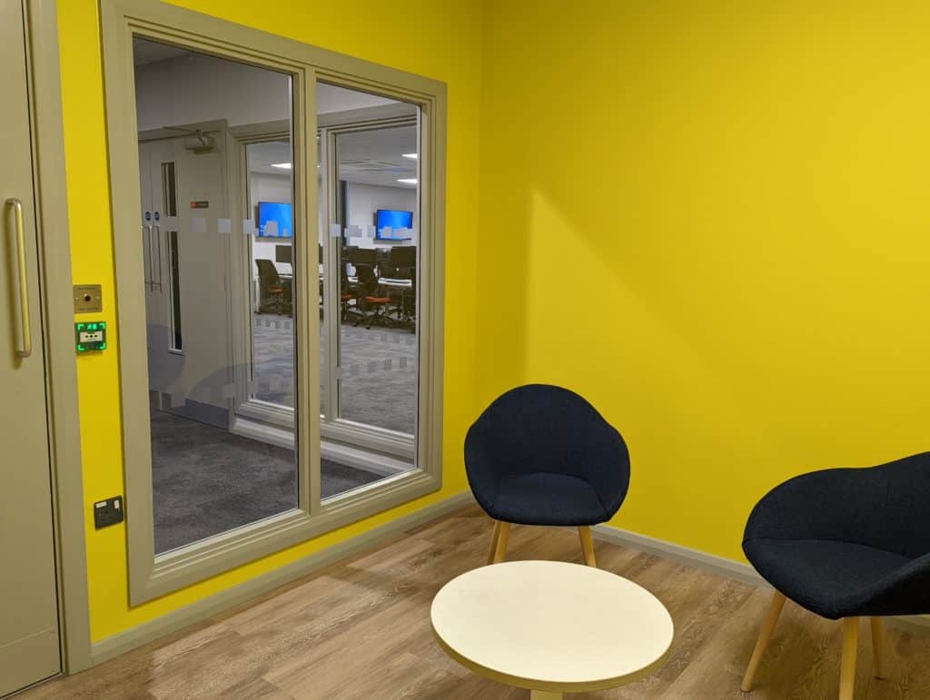 Inside the modular unit created for Hull University. Yellow walls, seating area with two comfy black chairs and a small round table.
