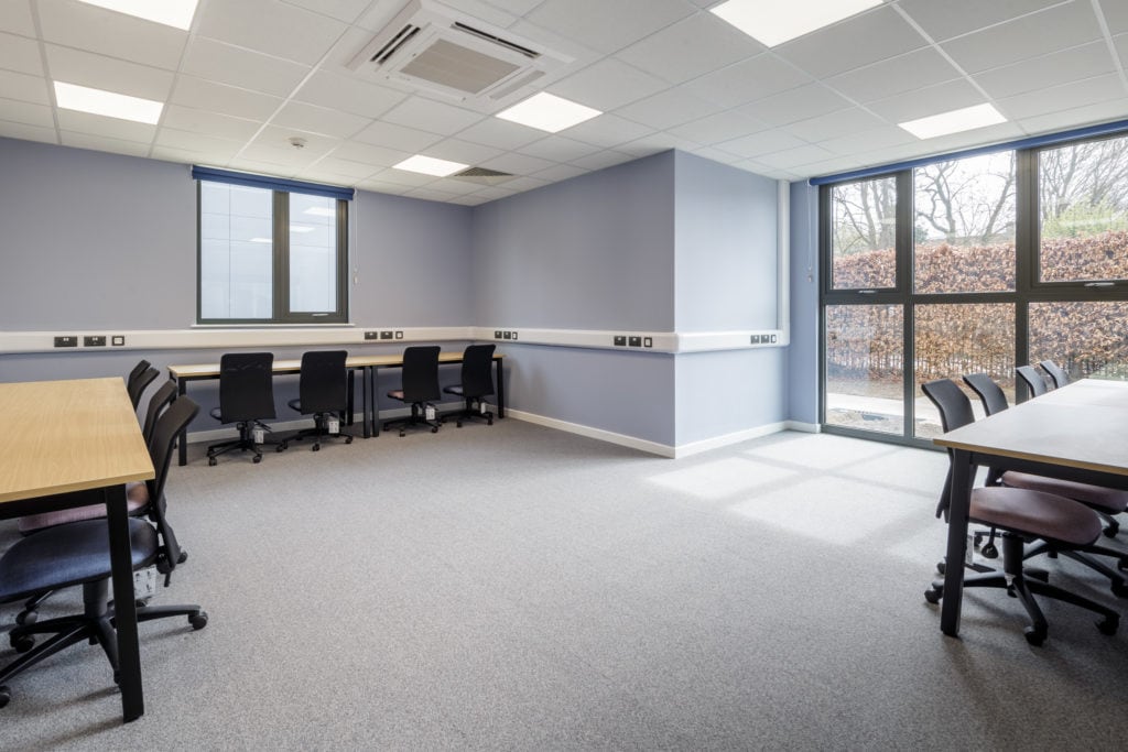 Inside a modular classroom, natural lighting. Desks with computer chairs, and a full length window next to the wall. 