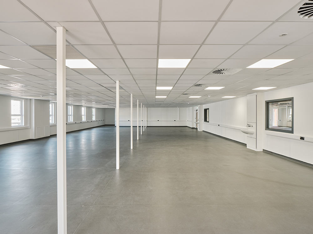 Large empty room inside temporary modular building. White walls, with white ceiling tiles and LED lighting. 