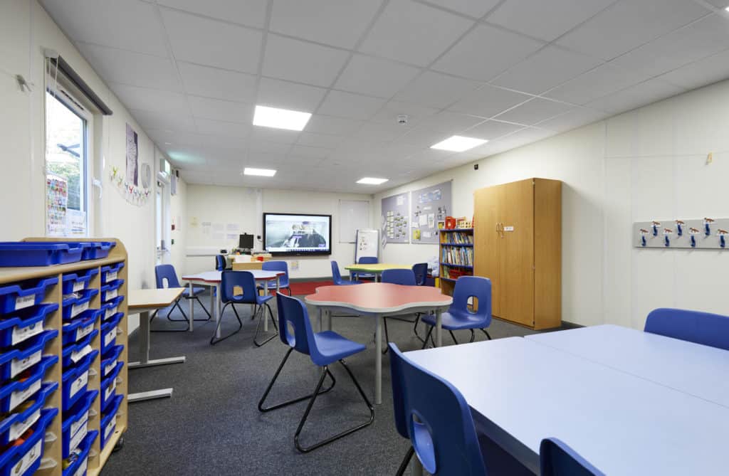 Inside temporary classrooms. 3 tables surrounded by chairs, interactive whiteboard on the wall. Bookcase, cabinet. Colourful room. Draw filles with student's trays. 