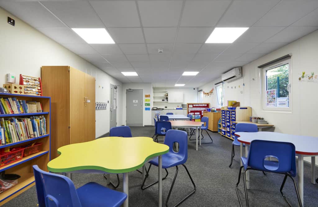 Inside modular classroom, colourful tables with chairs. Bookcase, cupboard, student's trays. 