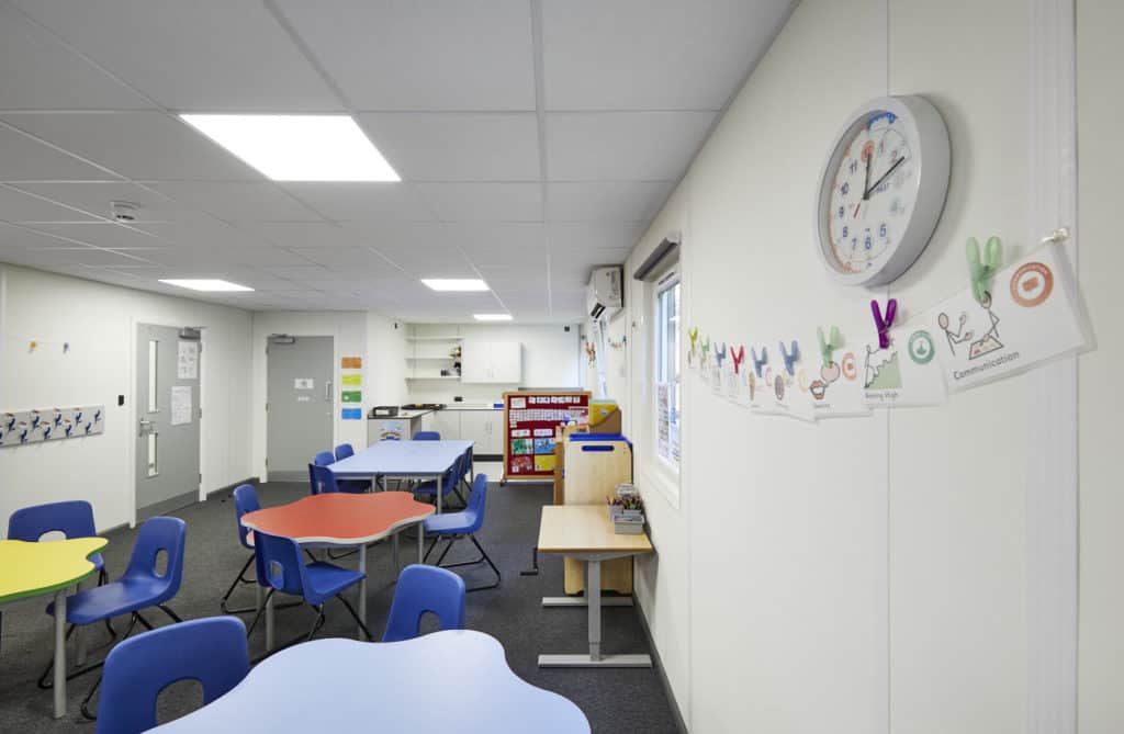 Inside temporary classroom. Colourful tables, with chairs. Clock on the wall, with learning materials hung up. 