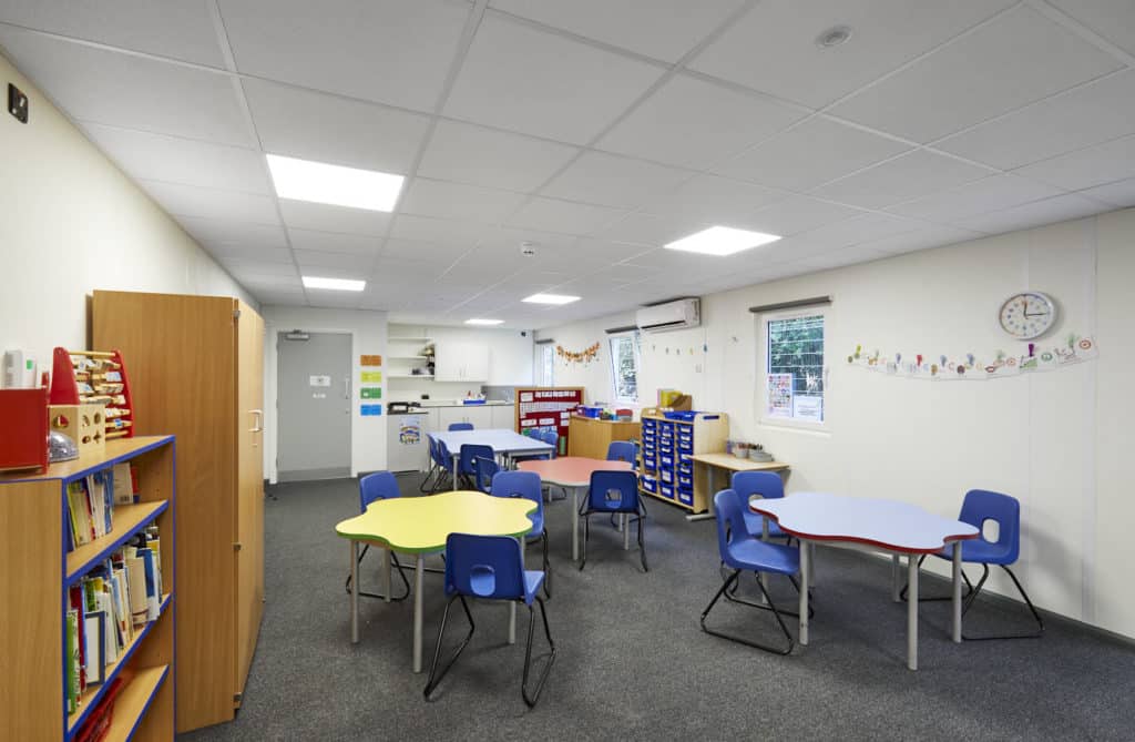 Inside temporary classrooms. 3 tables surrounded by chairs, interactive whiteboard on the wall. Bookcase, cabinet. Colourful room. Draw filles with student's trays. 