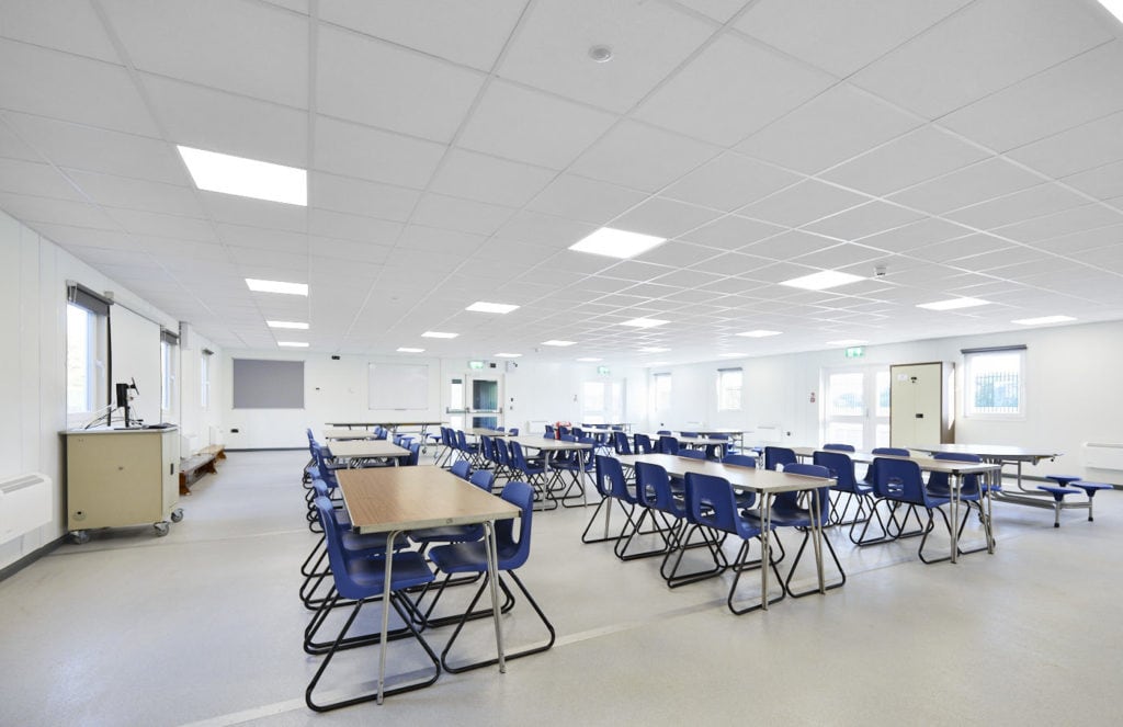 Inside temporary classroom. Several rectangle tables surrounded by blue plastic chairs. teachers desk at the front. 