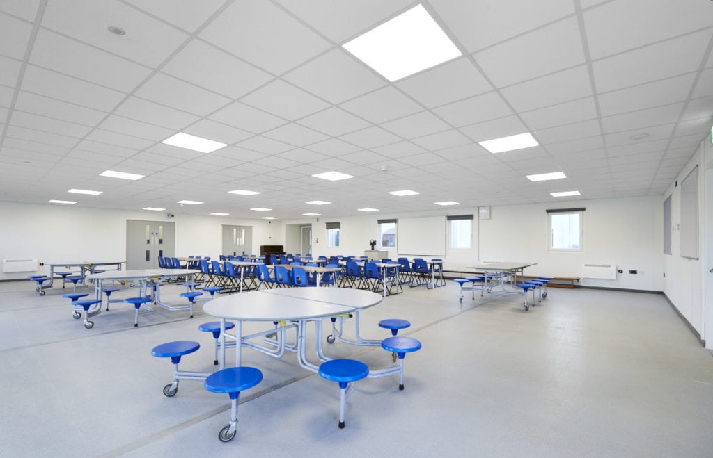 Cafeteria area inside temporary school building built for Bradsfield Academy. Several round tables surrounded by bleu stools. 
