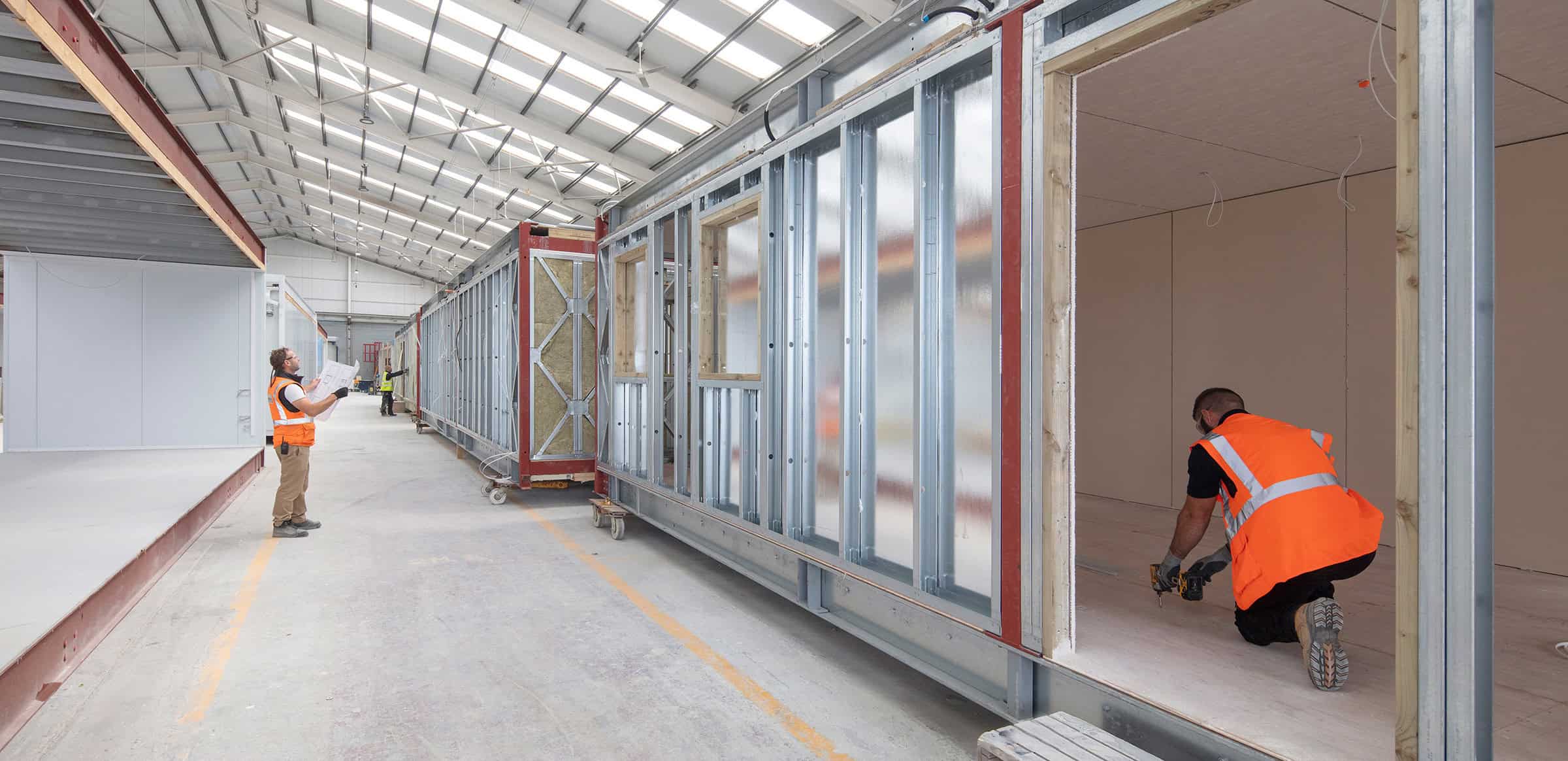 Temporary modular buildings being created offsite in a factory. 2 workers, one inside a modular unit drilling the floor, one stood looking at the plans. 
