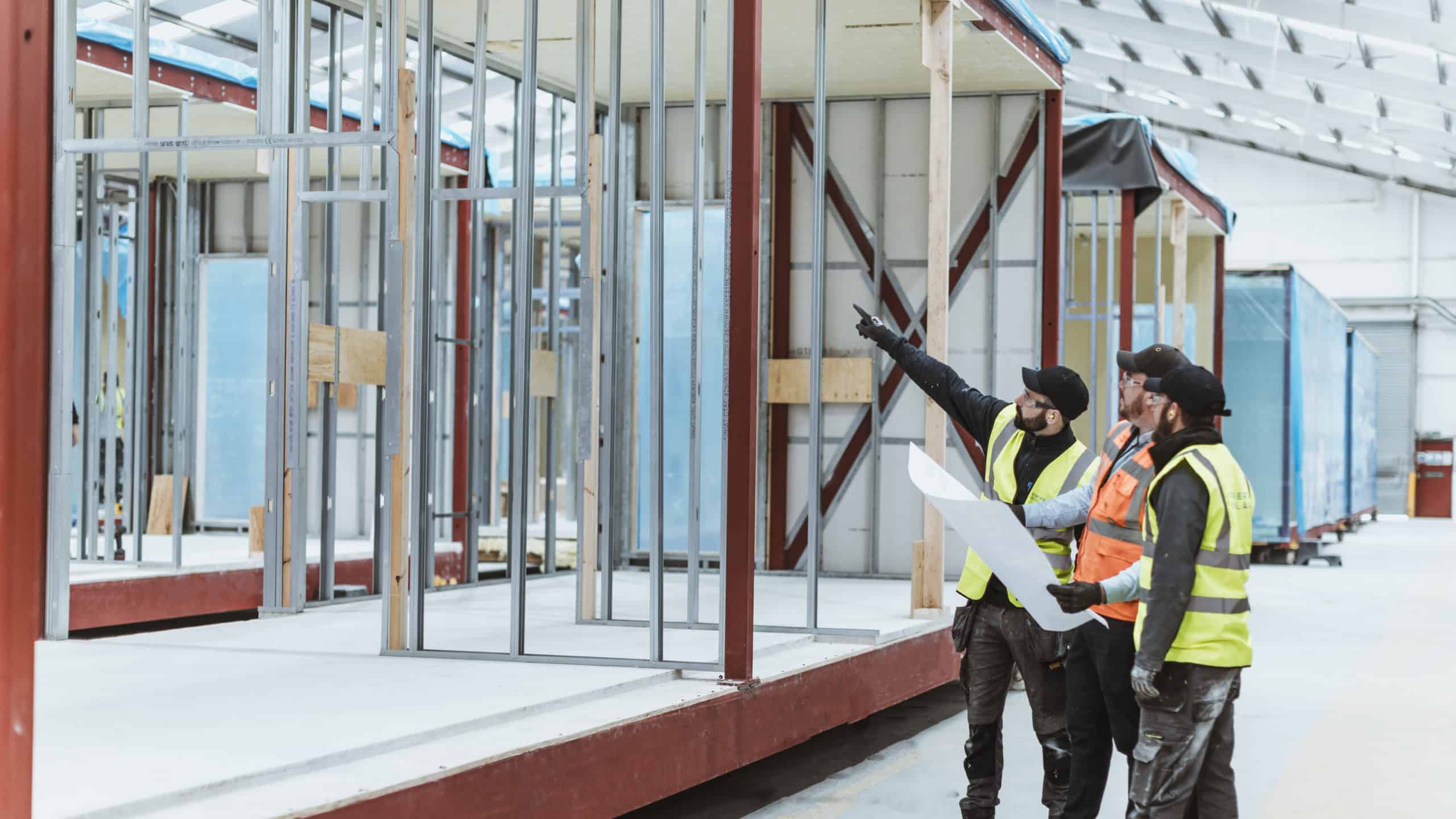 View inside a modular construction companies offsite factory. 3 workers in high vis vests holding a plan and pointing at a in-construction modular building. 