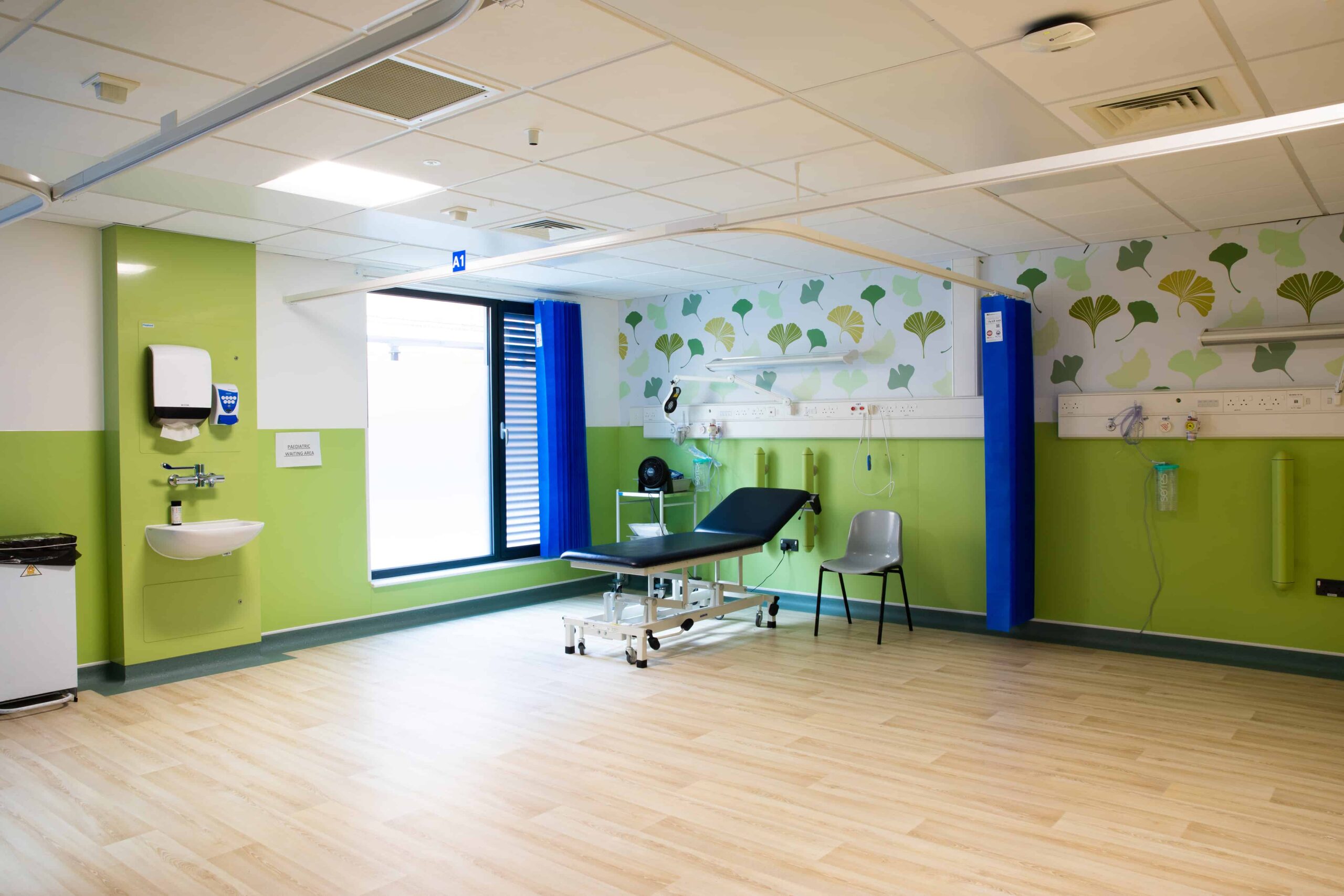 Inside view of a modular healthcare building, lime green walls, hospital bed, sink, grey chair, window. 