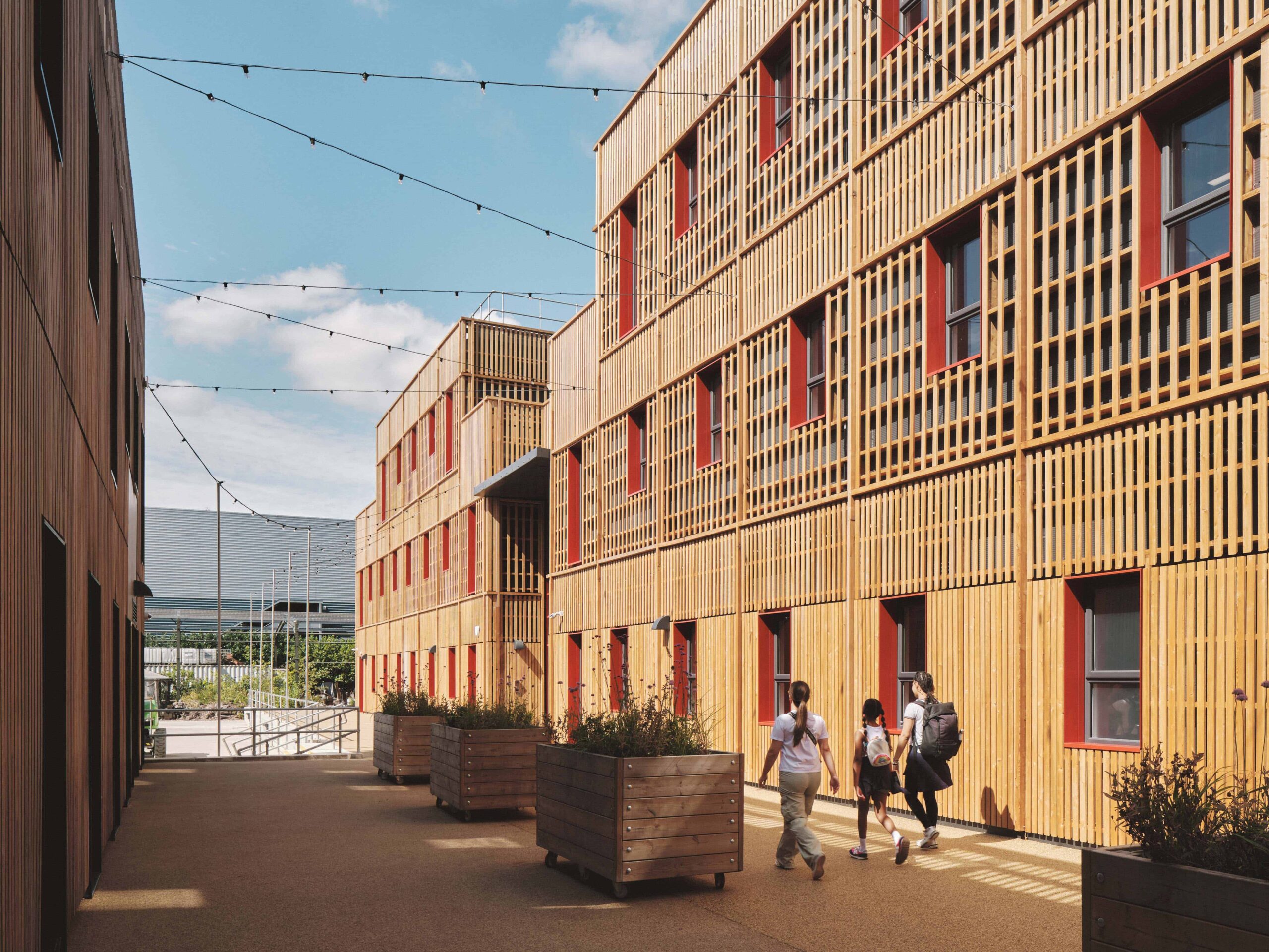 A family walking on the campus of The Paper yard, temporary modular structure. Multiple large buildings, with red window panes and wooden panelling on the walls. 