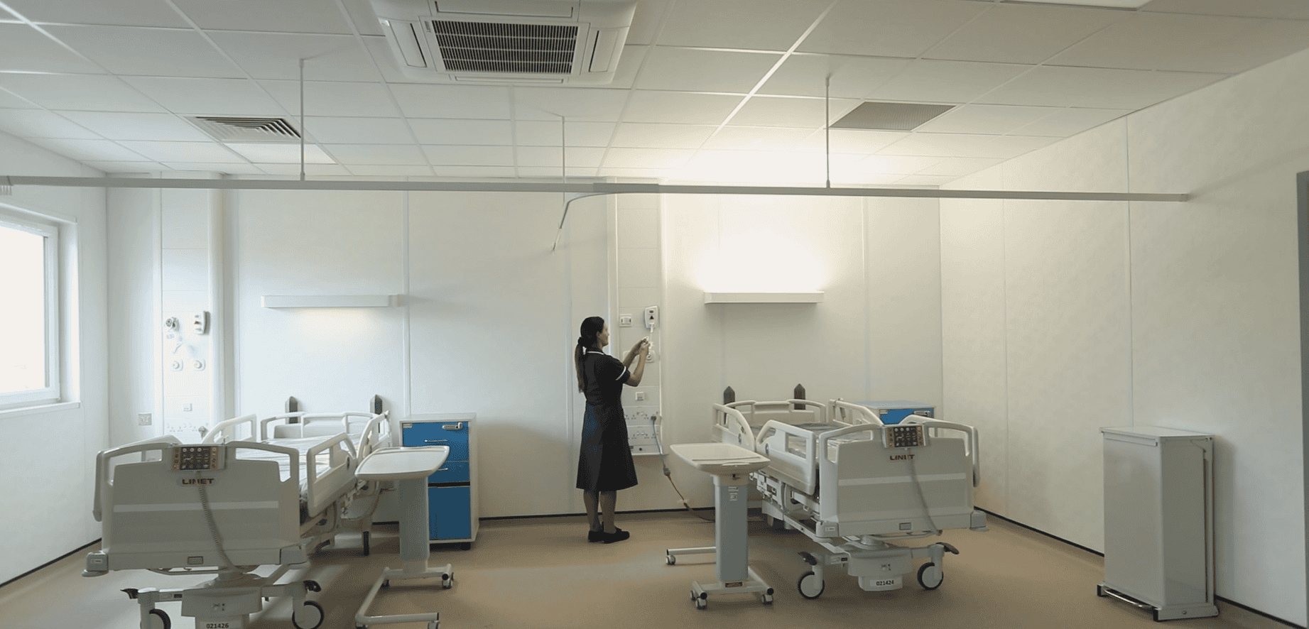 Hospital room inside a modular healthcare building. A nurse checking some machinery on the wall, two hosptial beds with desks. 
