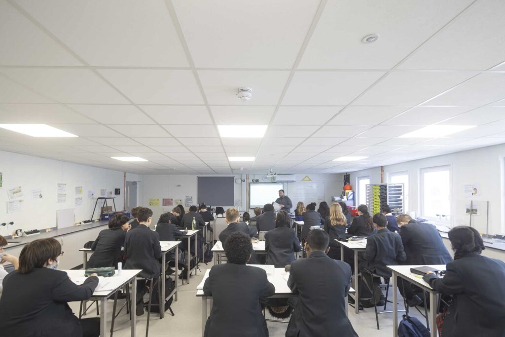 Inside Temporary classroom, filled with students wearing black blazers, sat at desks. Teacher at the front of the room standing next to the interactive whiteboard.