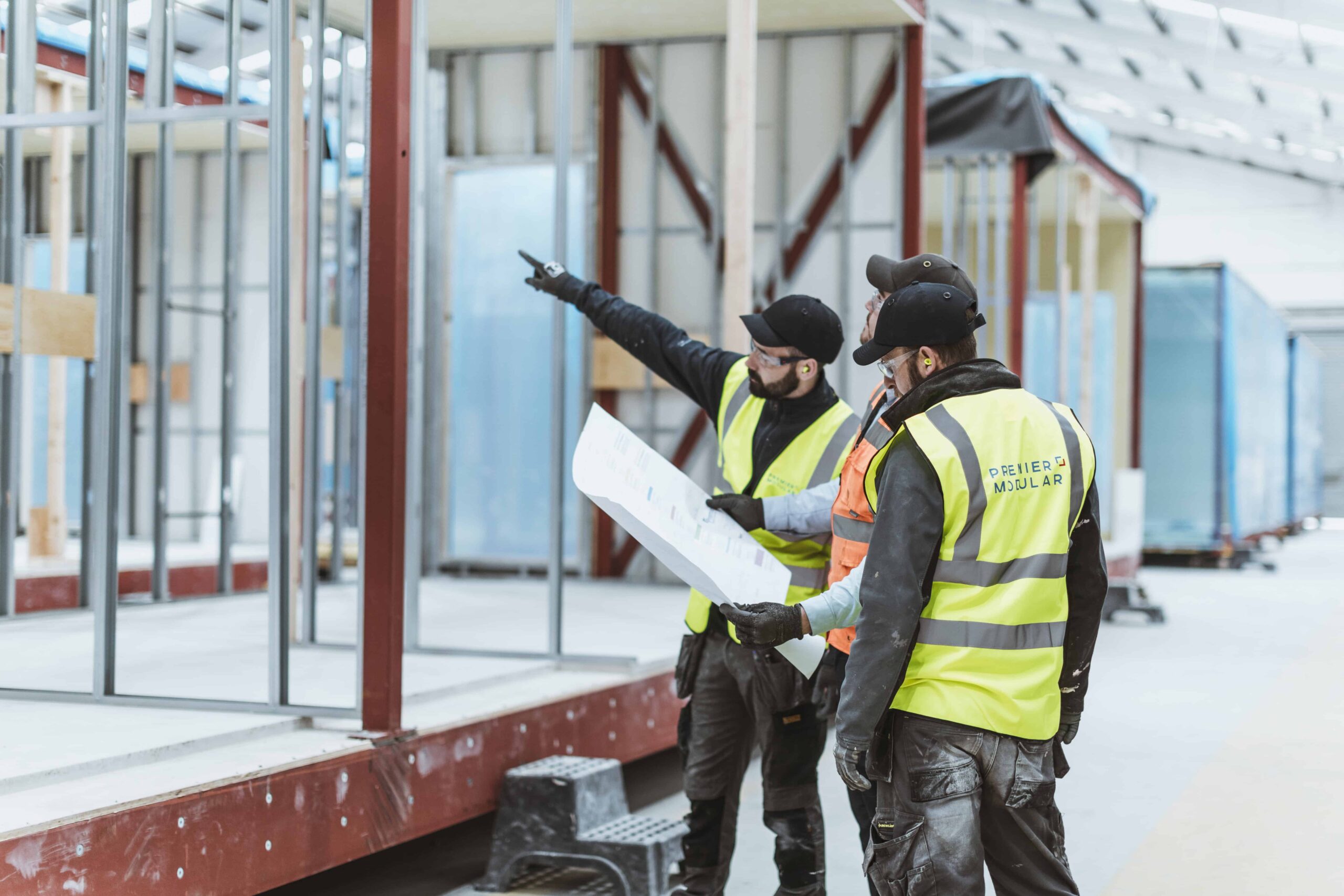 Three permier modular construction workers wearing high vis vests. Based in the offsite construction factory, surrounding some plans. One of them is pointing towards an unfinished modular building. 