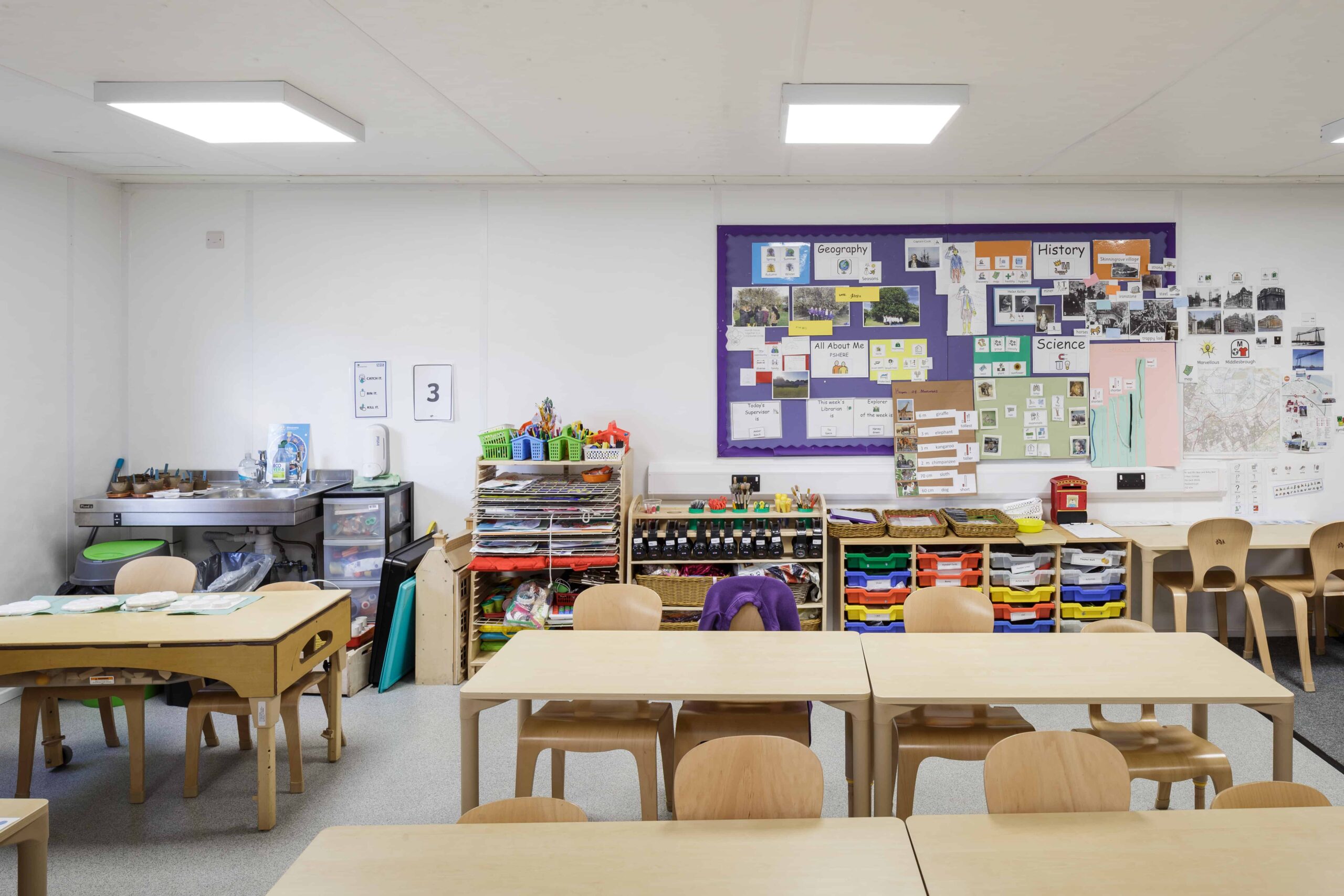 Inside temporary classroom, wooden desks and chairs, with student drawers and a cork board with work hung up. 