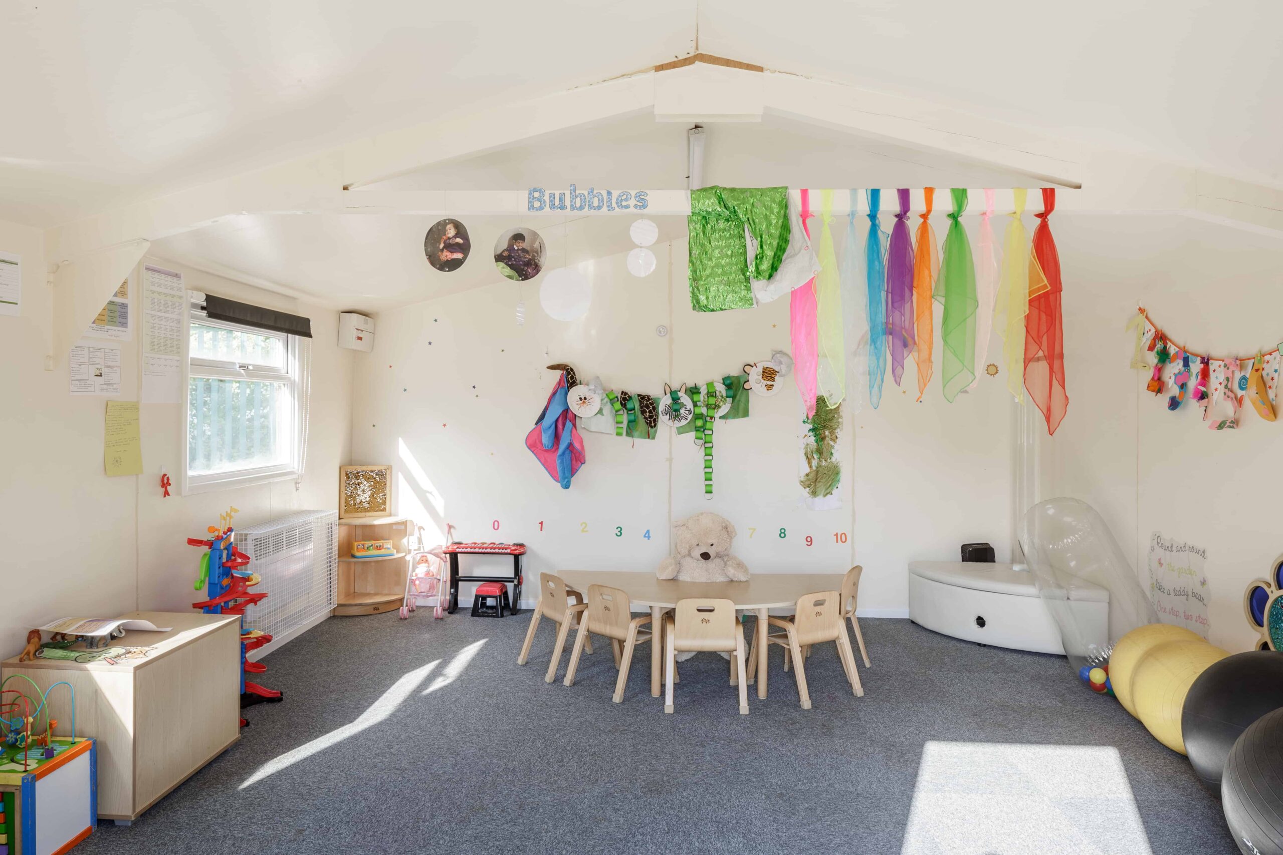 Temporary classroom for small children. Small desk with 5 chairs round it, with a teddy bear on the table. Colourful cloth hung on the beams, with toys around the walls. 