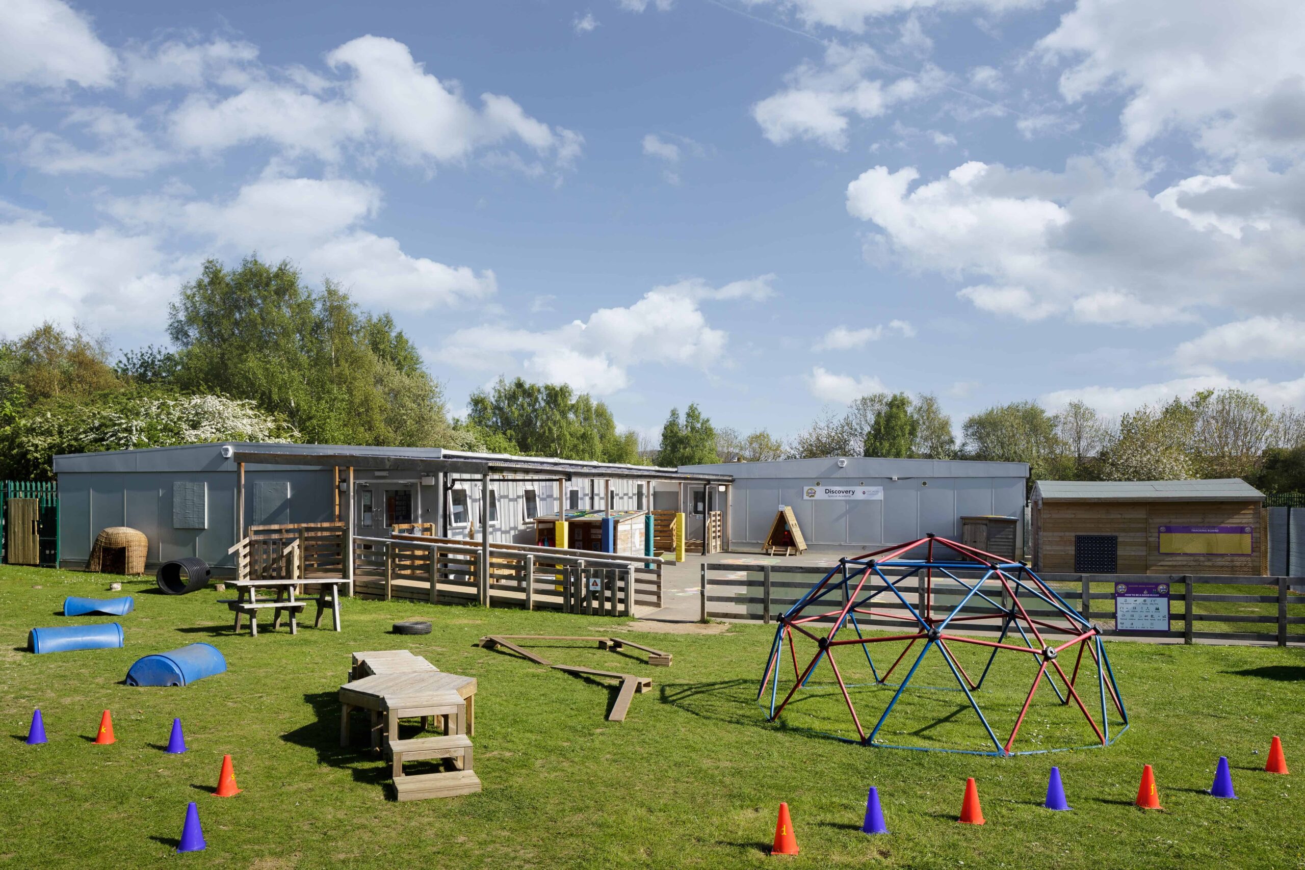 Full view of the outside of the temporary classrooms. play area on grass, with small climbing frame and cones. 