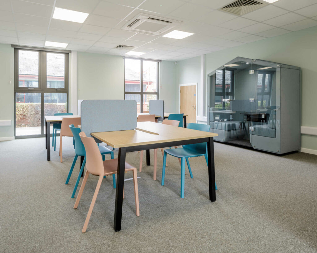 Inside a modular classroom. Wooden tables and chairs, with dividers between desks. Also featuring a small house with seating and a table for meetings.