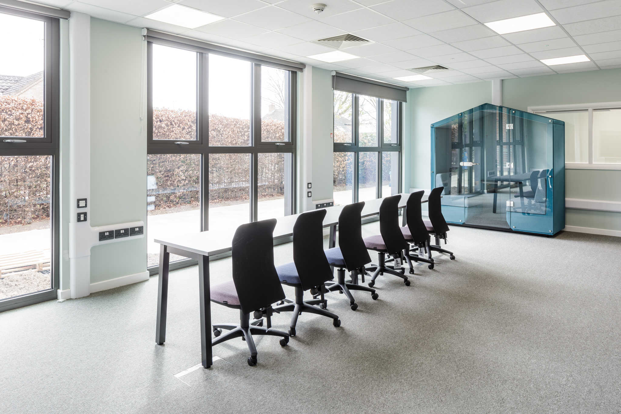 Open space modular building. Table and chairs in a room filled with natural lighting. Glass house booth for meetings. Lots of windows. 