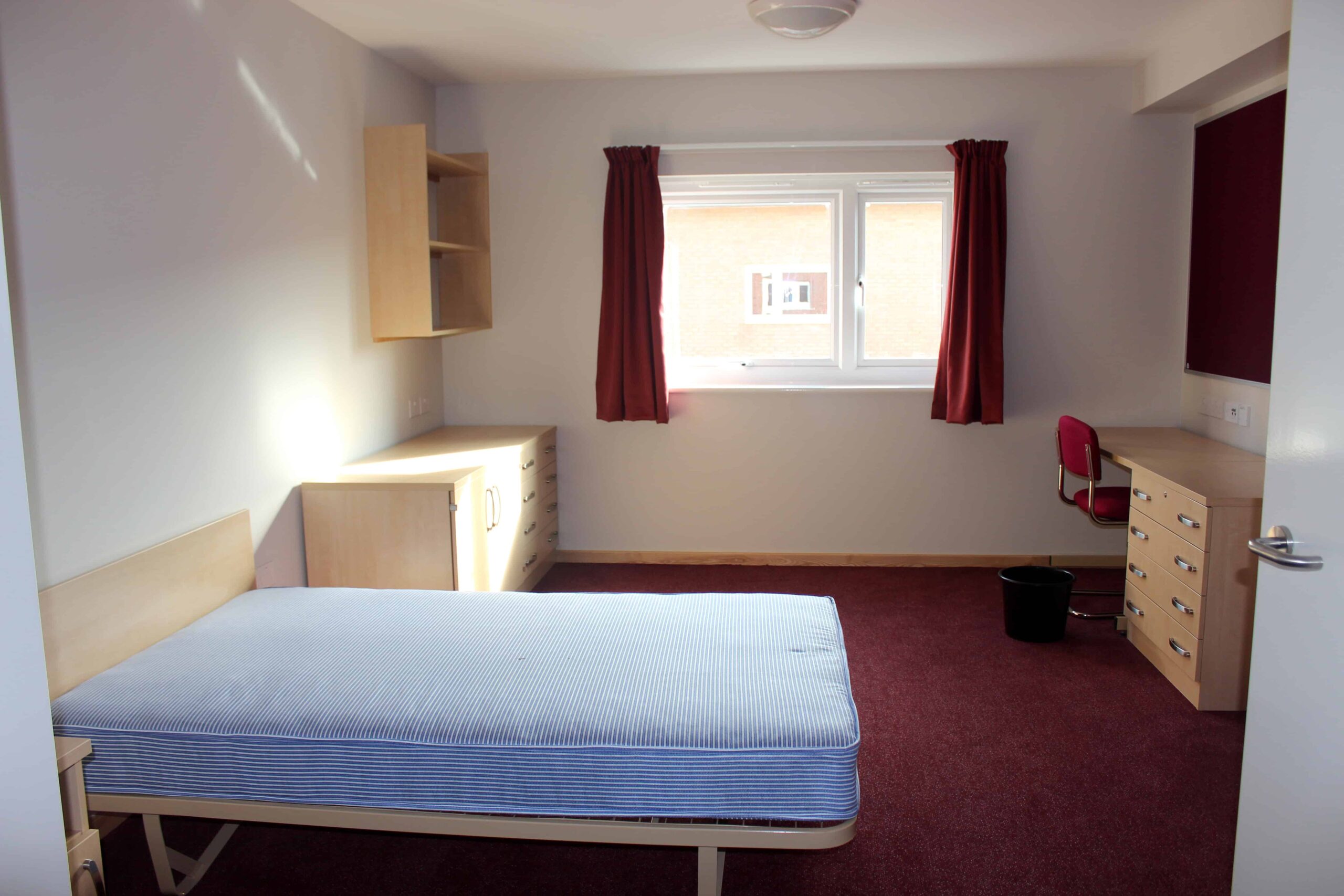 Single room inside modular accomodation complex. Single bed, desk and chair, drawers, shelves, and a window with red curtains. 